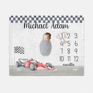 May include: A white baby blanket with a checkered border and a red race car illustration. The blanket also features a trophy, the numbers 1-12, and the text "Michael Adam" and "months".