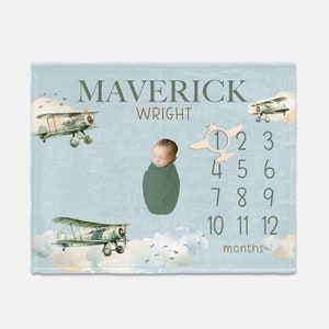 May include: A blue baby milestone blanket with a watercolor sky and clouds. There are three vintage airplanes and a baby boy in the center. The blanket has the name "Maverick Wright" and numbers 1-12 for each month.