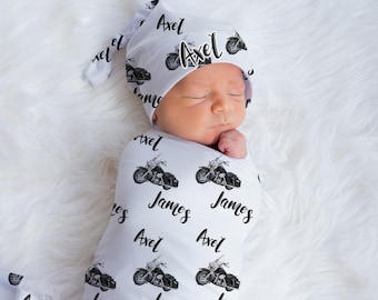May include: A white swaddle blanket with a black and white motorcycle print and the name "Axel" repeated throughout. A baby is swaddled in the blanket and wearing a matching hat.