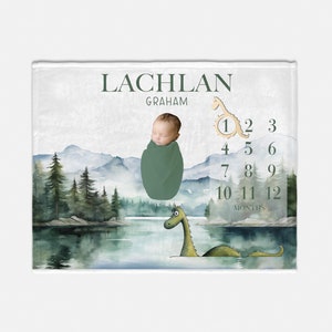 May include: A personalized baby milestone blanket featuring a watercolor landscape with mountains, trees, and a lake. The blanket includes the name "LACHLAN GRAHAM" and a dinosaur graphic. A newborn baby is swaddled in a green blanket.