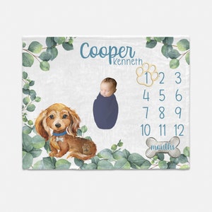May include: A white baby blanket with a watercolor design of eucalyptus leaves and a dachshund. The blanket has a blue and gold paw print design with numbers 1-12 for a baby's monthly milestones. The text "Cooper Kenneth" and "months" are printed on the blanket.