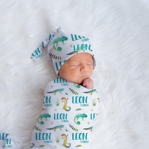 May include: A white swaddle blanket with a green and yellow chameleon print and the name "LEON" repeated throughout. A baby is swaddled in the blanket and is wearing a matching hat.