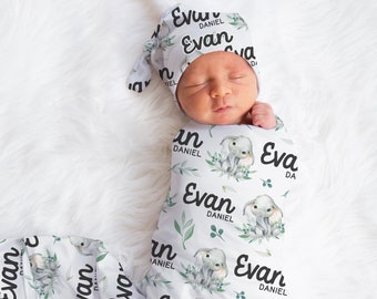 May include: A white swaddle blanket with a repeating pattern of green leaves, gray elephants, and the name "Evan Daniel". A baby is swaddled in the blanket and wearing a matching hat.