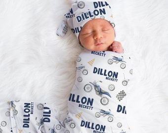 May include: A white swaddle blanket with a blue and white pattern of motorcycles and the name "Dillon Beckett" repeated throughout. A baby is wearing the swaddle and a matching hat.
