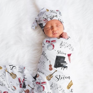 May include: A white swaddle blanket with a musical theme. The blanket features a variety of musical instruments, including a piano, guitar, drums, and a saxophone. The blanket is decorated with musical notes and the name "Steven" is repeated throughout the design. A baby is swaddled in the blanket and wearing a matching hat.