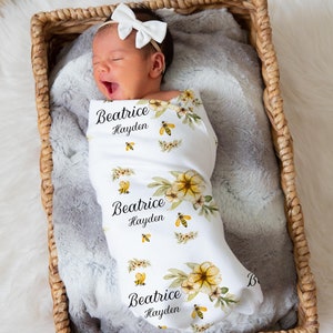 May include: A white swaddle blanket with a floral and bee design. The blanket has the names "Beatrice Hayden" printed on it. A baby is wrapped in the blanket and lying in a wicker basket.