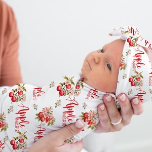 May include: A white swaddle with a red apple and floral pattern and the text "Apple Marie" repeated. A baby is wearing the swaddle and a matching headband with the same pattern and text.