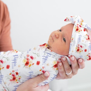 May include: A white swaddle with a floral pattern and the name "Poppy" repeated in purple text. A baby is wrapped in the swaddle and wearing a matching headband with the same floral pattern and text.