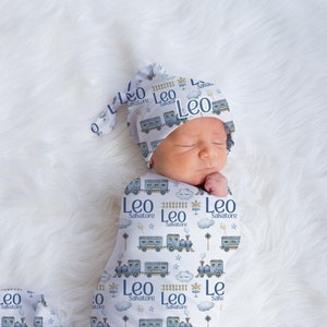 May include: A white baby swaddle with a blue train pattern and the name "Leo Salvatore" repeated throughout. The swaddle is wrapped around a baby wearing a matching blue train hat.