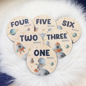 May include: A set of six wooden monthly milestone discs. Each disc features a space-themed illustration with a rocket, planets, and an astronaut. The discs are labeled with the months "ONE" through "SIX".