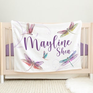May include: White fleece blanket with a purple floral design and colorful dragonfly illustrations. The blanket has the name "Mayline Shea" written in purple cursive.