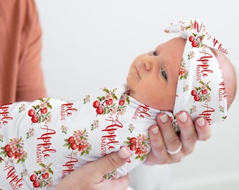 May include: A white swaddle with a red apple and floral pattern and the text "Apple Marie" repeated. A baby is wearing the swaddle and a matching headband with the same pattern and text.