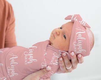 May include: A pink swaddle blanket with the name "Aaliyah Grace" printed on it. A baby is wrapped in the blanket and wearing a matching pink headband with the same name.