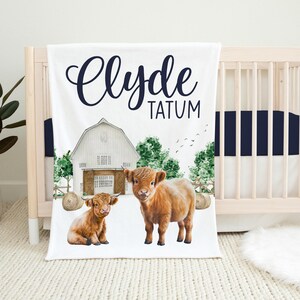 Op de afbeelding: Een witte babydeken met een rode schuur, groene bomen en twee bruine Highland-koeien. De deken heeft de naam "Clyde Tatum" in zwarte cursieve letters.