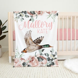 May include: A white baby blanket with a pink floral design and a flying duck. The blanket has the name "Mallory Kate" written in pink cursive.