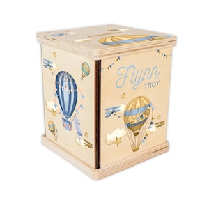 May include: A square wooden money box featuring light wood grain and printed illustrations of blue and tan hot air balloons, small airplanes, clouds, and stars. The name Flynn Troy is printed in blue script on the front panel with a coin slot.
