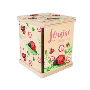 May include: A wooden box with a ladybug design and the name "Louise Josephine" written on the side. The box is decorated with red ladybugs, green leaves, and pink flowers.