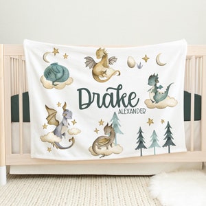 May include: White baby blanket with a green, blue, and gold dragon theme. The blanket features five dragons, stars, and the name "Drake Alexander".
