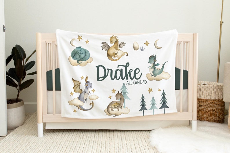 May include: White baby blanket with a green, blue, and gold dragon theme. The blanket features five dragons, stars, and the name "Drake Alexander".