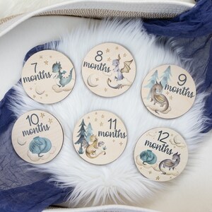 May include: Set of six wooden monthly milestone discs. Each disc features a whimsical dragon illustration, the month number, and the word "months". The discs are decorated with stars, clouds, and trees.