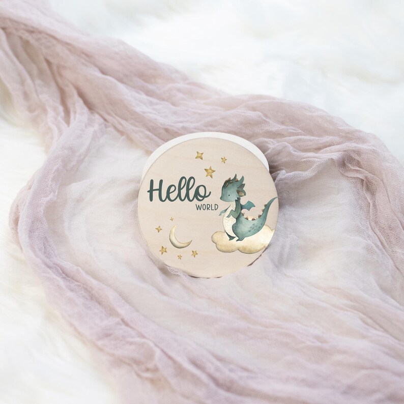 May include: A round wooden sign with the words "Hello World" in green script. A watercolor illustration of a teal dragon on a cloud is featured. Gold stars and a crescent moon surround the text and image. The sign is on a light pink fabric.
