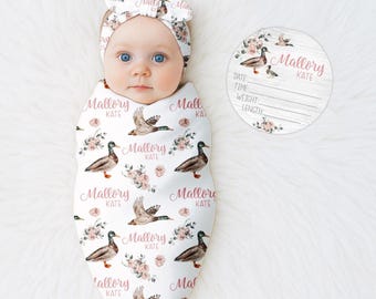 Op de afbeelding: Een baby gewikkeld in een witte deken en met een bijpassende hoofdband, beide versierd met een eenden- en bloemenpatroon. De deken en hoofdband zijn bedrukt met de naam "Mallory Kate". Een ronde houten plaat met hetzelfde ontwerp is ook aanwezig.