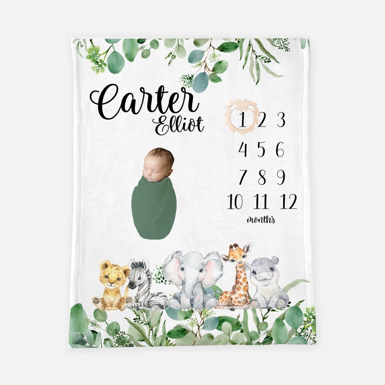 May include: A white baby blanket with a green leafy border. The blanket features a baby boy in a green swaddle, a lion, zebra, elephant, giraffe, and hippopotamus. The blanket also includes the text "Carter Elliot" and a number chart for tracking a baby's growth from 1 to 12 months.