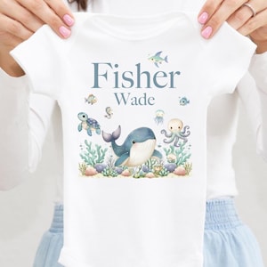 May include: White baby bodysuit with a blue and watercolor sea life design. The design includes a whale, octopus, turtle, and fish. The name "Fisher Wade" is printed in blue script above the sea life illustrations.