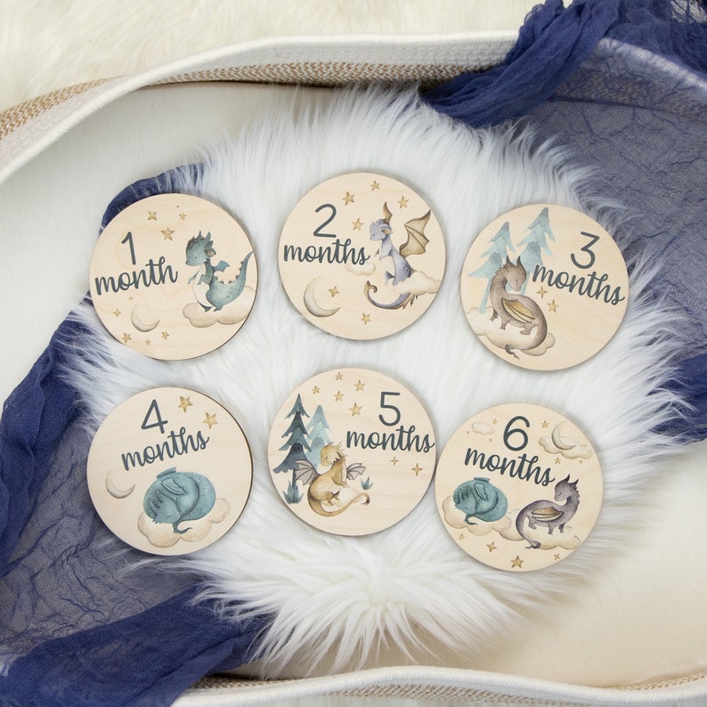 May include: Set of six wooden monthly milestone discs. Each disc features a different watercolor dragon illustration with the month number and the word "months". The discs are light brown with a soft, whimsical design.