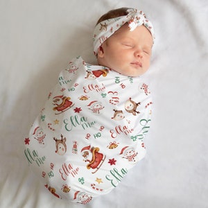 K&ouml;nnte beinhalten: Eine wei&szlig;e Wickeldecke mit Weihnachtsmotiv, die ein rot-gr&uuml;nes Muster aus Weihnachtsmann, Rentier, Schneem&auml;nnern und Geschenken zeigt. Die Decke tr&auml;gt den Namen "Ella Mae", der sich &uuml;ber das gesamte Design wiederholt. Ein Baby ist in die Decke gewickelt und tr&auml;gt ein passendes Stirnband.