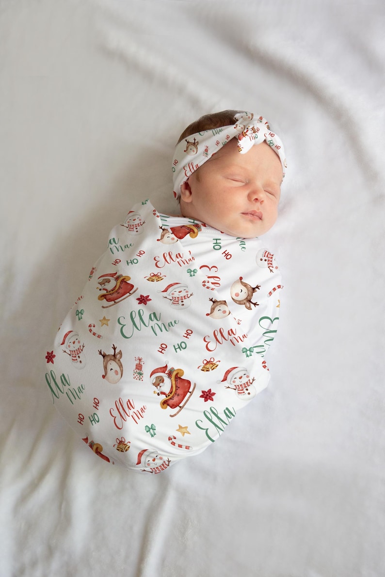 K&ouml;nnte beinhalten: Eine wei&szlig;e Wickeldecke mit Weihnachtsmotiv, die ein rot-gr&uuml;nes Muster aus Weihnachtsmann, Rentier, Schneem&auml;nnern und Geschenken zeigt. Die Decke tr&auml;gt den Namen "Ella Mae", der sich &uuml;ber das gesamte Design wiederholt. Ein Baby ist in die Decke gewickelt und tr&auml;gt ein passendes Stirnband.