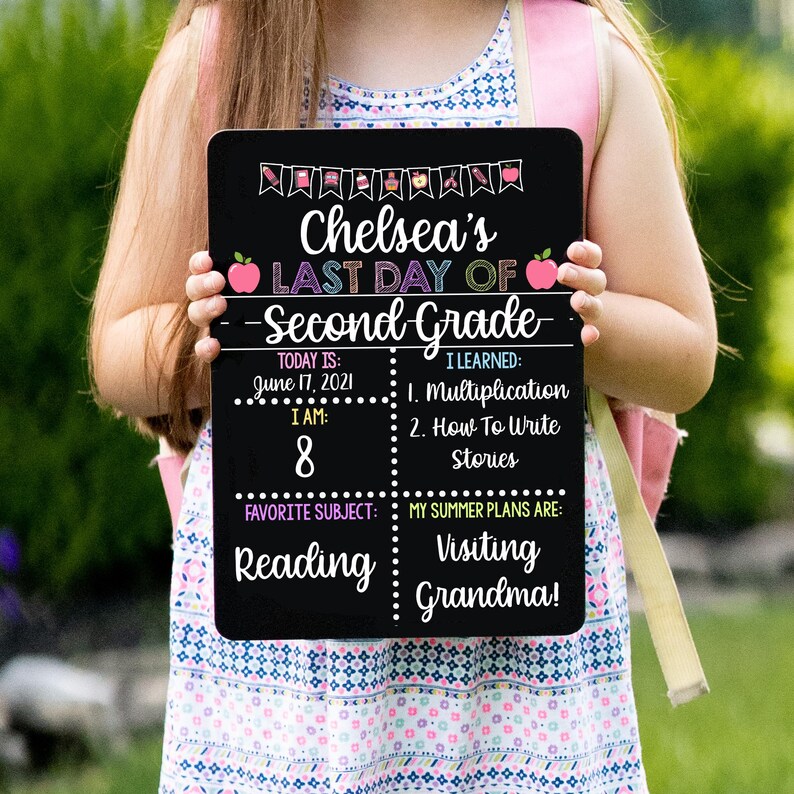 May include: Black chalkboard sign with the text "Chelsea's Last Day of Second Grade". The sign has a pink apple graphic, a calendar with the date June 17, 2021, and the number 8. It also includes the text "I Learned: Multiplication, How To Write Stories", "Favorite Subject: Reading", and "My Summer Plans Are: Visiting Grandma!"