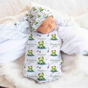 May include: A white swaddle with a green frog pattern and the name "Frederic Banks" repeated throughout. A matching white hat with the same pattern and name is on the baby's head.