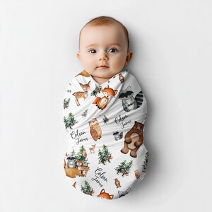 May include: A white swaddle blanket with a woodland animal print. The blanket features foxes, owls, deer, raccoons, and bears. The name "Colson James" is repeated throughout the design.