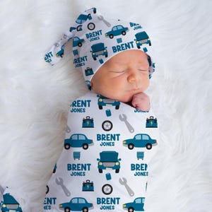 May include: A white swaddle blanket with a blue car and wrench pattern. The blanket has the name "Brent Jones" repeated throughout the pattern. A matching blue and white hat with the name "Brent Jones" is on top of the blanket.