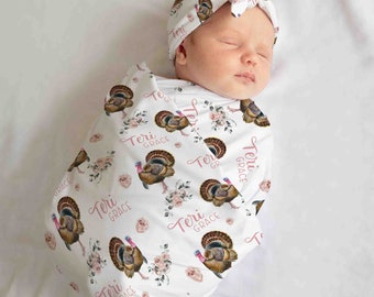 Op de afbeelding: Een slapende pasgeboren baby gewikkeld in een witte deken en bijpassende hoofdband. De deken heeft een patroon van kalkoenen, roze bloemen en de naam "Teri Grace". De baby slaapt vredig.