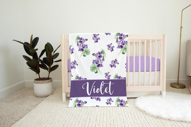 May include: A white baby blanket with a purple floral pattern and the name "Violet" printed in white on a purple band.