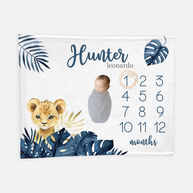 K&ouml;nnte beinhalten: Eine wei&szlig;e Baby-Meilenstein-Decke mit einem blauen und goldenen Dschungel-Motiv. Die Decke zeigt einen L&ouml;wen, tropische Bl&auml;tter und eine Zahlentabelle, um das Wachstum eines Babys zu verfolgen. Der Text "Hunter Leonardo" ist oben auf der Decke aufgedruckt.