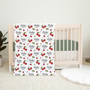 K&ouml;nnte beinhalten: Wei&szlig;e Babydecke mit einem sich wiederholenden Muster aus roten Kirschen und dem Text "Ruby Cait" in schwarzer Kursivschrift.