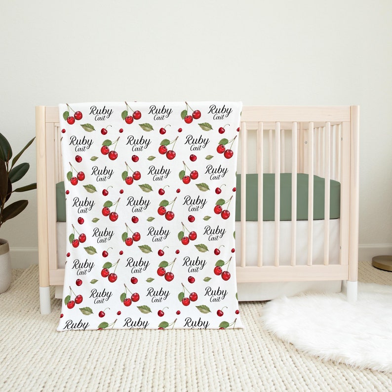 K&ouml;nnte beinhalten: Wei&szlig;e Babydecke mit einem sich wiederholenden Muster aus roten Kirschen und dem Text "Ruby Cait" in schwarzer Kursivschrift.