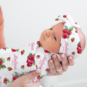 May include: A white swaddle blanket with a raspberry print and the name "Malina Marie" written in pink. A matching headband with the same print and name is on the baby's head.