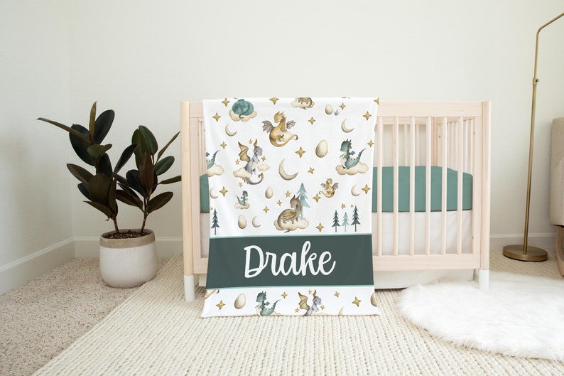 May include: A white baby blanket with a green border and a repeating pattern of dragons, stars, and moons. The blanket has the name "Drake" written in white on a green background.