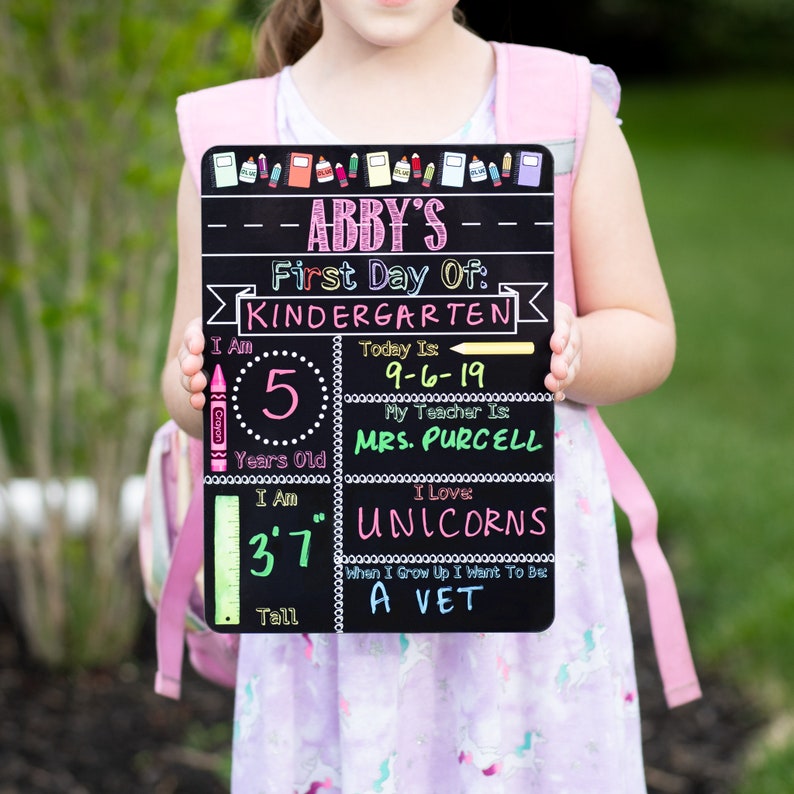 May include: A black chalkboard sign with the text "Abby's First Day Of: Kindergarten" and details about the child's age, height, and favorite things. The sign is held by a child wearing a pink dress and a pink backpack.