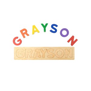 May include: A wooden name puzzle with the name "GRAYSON" in rainbow-colored letters. The letters are arched above a wooden base with the name carved into it. The puzzle is designed for children.