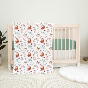 K&ouml;nnte beinhalten: Eine wei&szlig;e Babydecke mit einem sich wiederholenden Muster aus Weihnachtsmann in einem roten Schlitten, Rentieren, Zuckerstangen und Sternen. Der Text "Ella Mae" wird im gesamten Muster wiederholt.