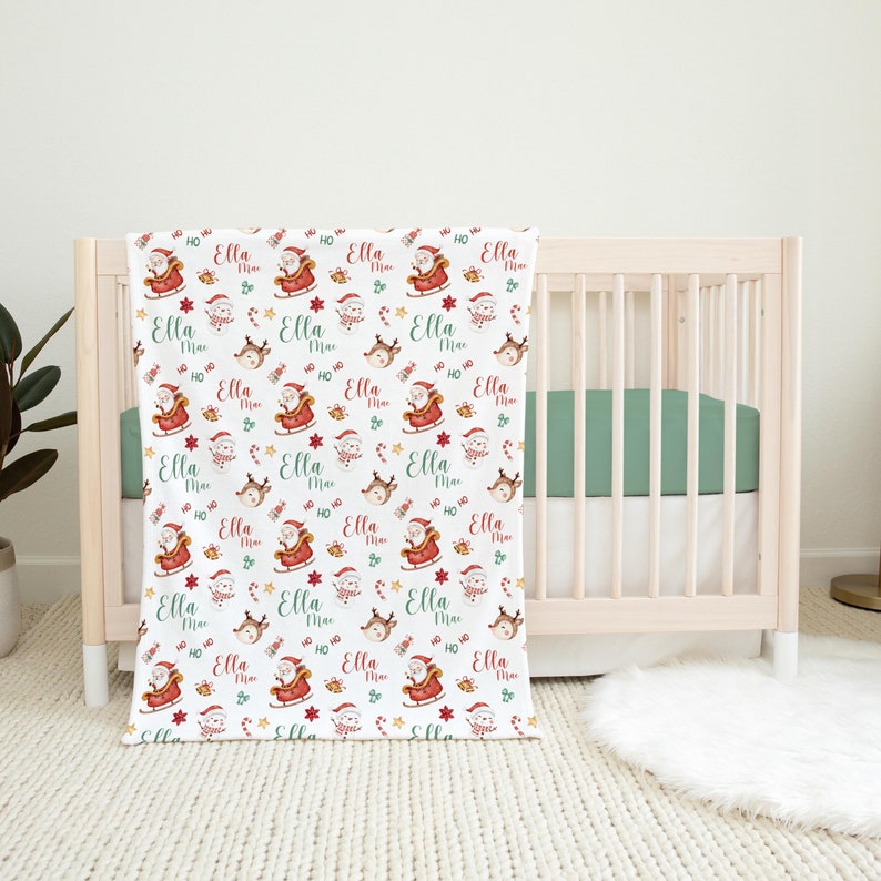 K&ouml;nnte beinhalten: Eine wei&szlig;e Babydecke mit einem sich wiederholenden Muster aus Weihnachtsmann in einem roten Schlitten, Rentieren, Zuckerstangen und Sternen. Der Text "Ella Mae" wird im gesamten Muster wiederholt.