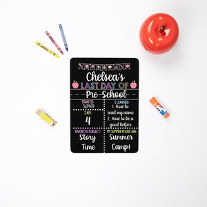 May include: Black chalkboard sign with the text "Chelsea's Last Day of Pre-School" with a red apple on each side of the text. The sign also includes the date "5/24/21", the age "4", favorite subject "Story Time", and summer plans "Summer Camp!"