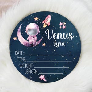 May include: A round, navy blue sign with a space theme. It features an astronaut on a crescent moon, a rocket, and stars. The sign includes lines for entering a baby's birth details, with the name "Venus Lyra" in elegant script.