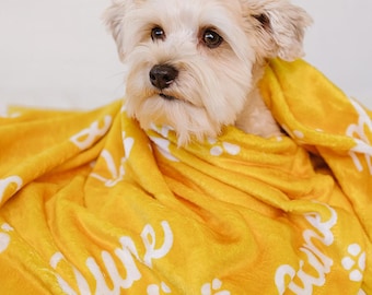May include: A small, light-colored dog peeks out from a bright yellow blanket. The blanket has the word "June" in a cursive font, along with white paw prints. The dog has a soft, fluffy coat and is looking towards the camera.