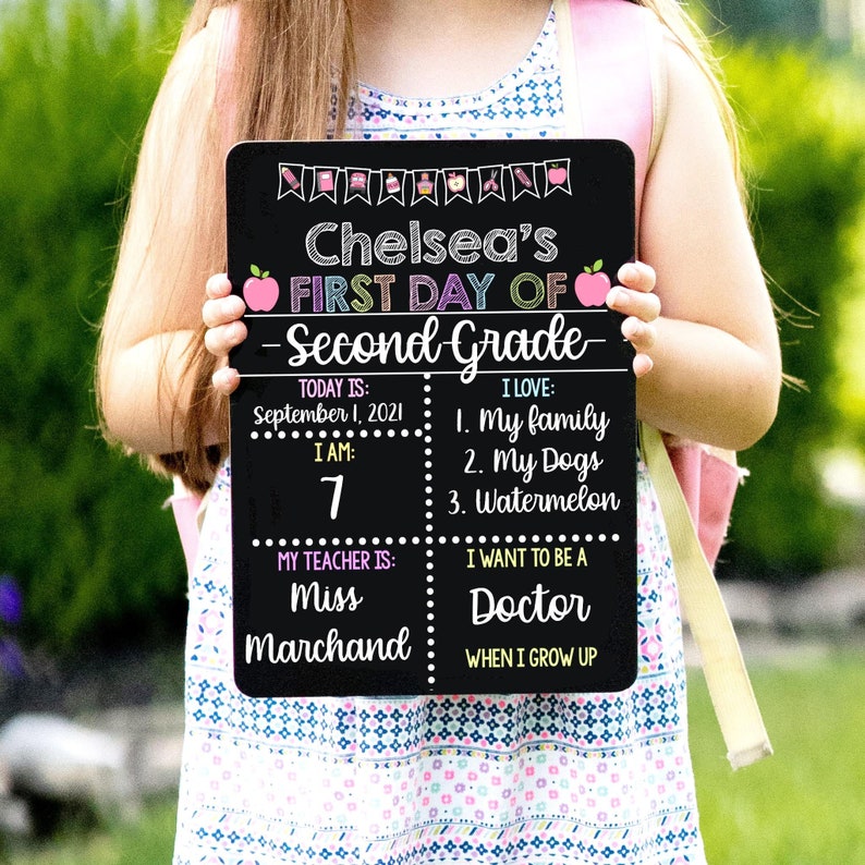 May include: A black chalkboard sign with the text "Chelsea's First Day of Second Grade". The sign has a pink apple on each side of the text. The sign is divided into four sections. The top left section says "Today is: September 1, 2021". The top right section says "I Love: 1. My family 2. My Dogs 3. Watermelon". The bottom left section says "I am: 7 My Teacher is: Miss Marchand". The bottom right section says "I want to be a Doctor When I grow up".